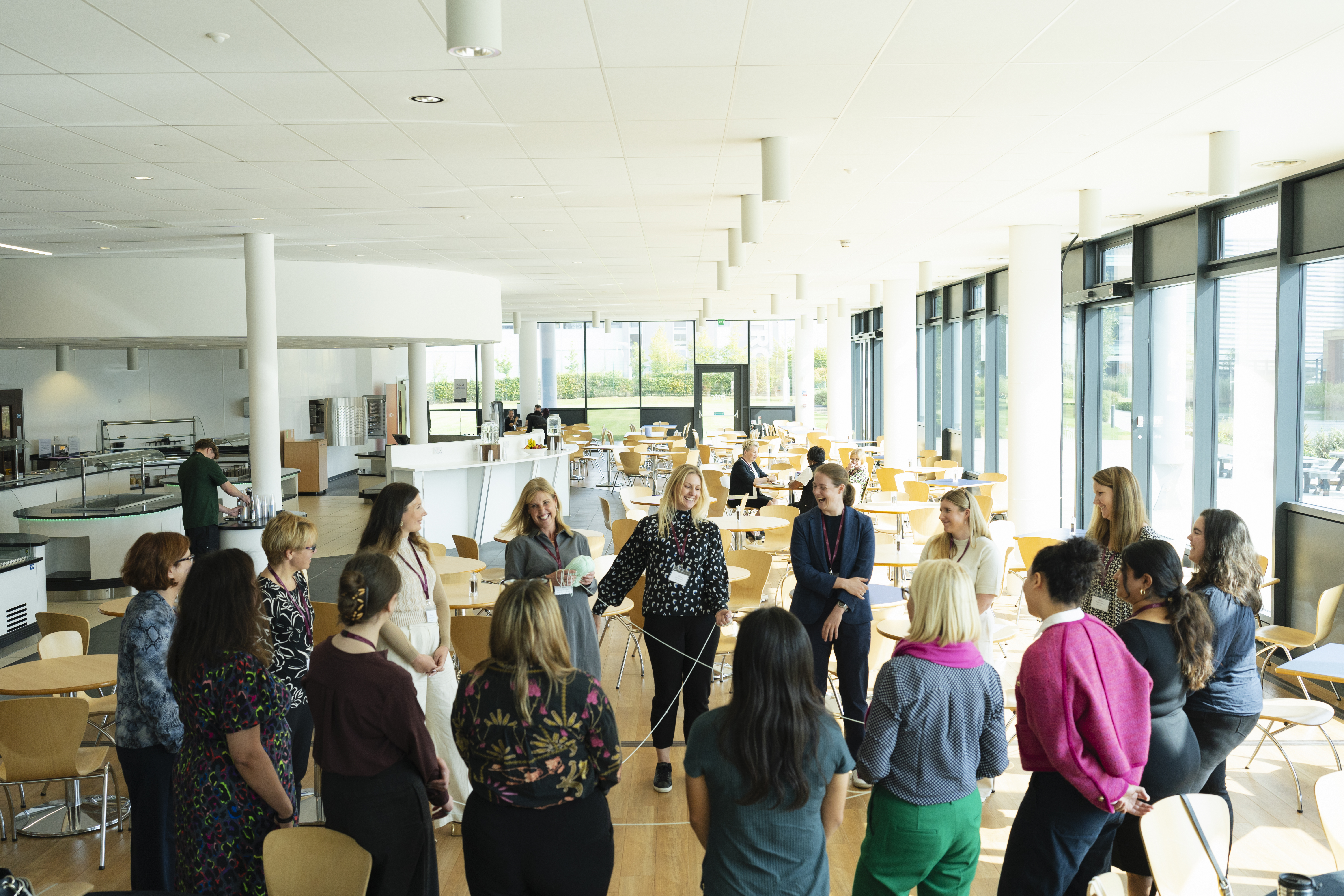 A group of women stand in a large circle in a bright, modern common room, participating in a networking or team-building activity. The room is filled with natural light from large windows.